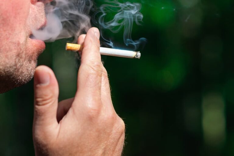 Pessoa fumando um cigarro, com foco no rosto e na mão segurando o cigarro aceso, liberando fumaça.